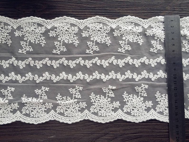 May include: Off-white lace fabric with floral and leaf patterns. The lace features a scalloped edge and is measured with a ruler marked in inches. The fabric is likely used for sewing, crafts, or decorative purposes.