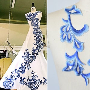 以下が含まれることがあります： 青い花の刺繍が施された白いドレス。刺繍は葉と花をモチーフにした繊細なデザインです。ドレスはノースリーブで、体にフィットするボディスです。スカートはボリュームがあり、流れるようなシルエットです。