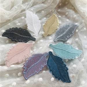 Peut inclure: Assortiment de sept patchs en tissu brodés en forme de feuilles dans différentes couleurs, notamment blanc, beige, gris, noir, rose, violet et bleu. Les patchs sont disposés en cercle sur un fond blanc.