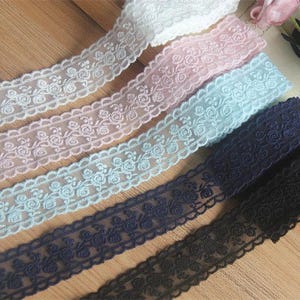 May include: A collection of five rolls of lace trim featuring a repeating floral rose pattern. The lace strips are available in white, light pink, light blue, dark blue, and black, each with scalloped edges and a delicate, semi-sheer mesh texture.