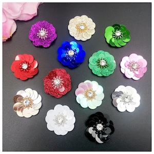 May include: A collection of sequined flower embellishments in various colors, including purple, gold, blue, green, red, and pink. Each flower has a central rhinestone. These decorative flowers are suitable for crafts and embellishments.