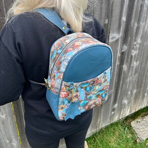 May include: A teal blue leather backpack with a colorful agate print. The backpack has two zippered compartments and a small leather tag.
