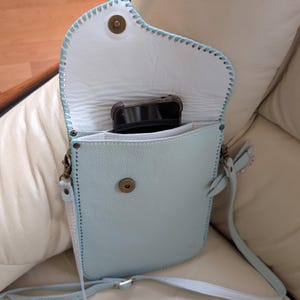 Puede incluir: Bolso bandolera de cuero azul claro con interior blanco. El bolso tiene un cierre de solapa con un broche magnético y una correa larga y ajustable. Se ve un smartphone dentro del bolso abierto. Los bordes están cosidos con hilo azul claro.