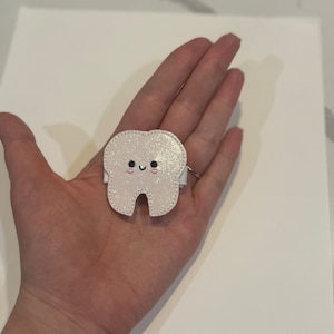 May include: A white glitter tooth with a pink and black smiling face. The tooth is a felt applique.