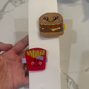 May include: Two felt food clips, one shaped like a hamburger with a smiling face and eyelashes, and the other shaped like a red french fry container with a smiling face and a purple bow.