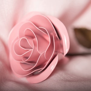 May include: A glossy pink rose with detailed, layered petals. The rose is set against a soft pink fabric background. The image highlights the rose's form and texture, creating a delicate and romantic feel.