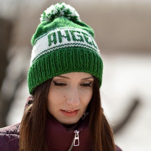 May include: A woman wearing a green and white knit beanie with a pom pom on top. The beanie has the word "WINGER" written on it in white.