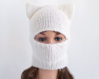 May include: White knitted cat ear beanie with a face mask. The beanie has a white knit with a textured pattern. The face mask covers the nose and mouth and has eye holes.