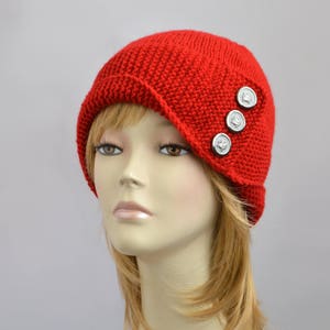 May include: A red knitted hat with a wide brim and silver buttons. The hat is angled on the side and has a simple design.