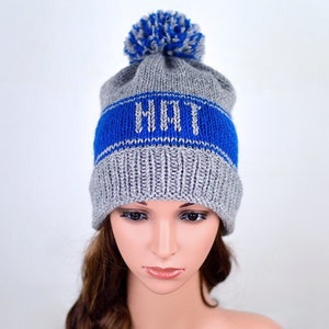 May include: Gray knit beanie with a blue stripe and the word "HAT" in white lettering. The beanie has a fluffy pom pom on top.