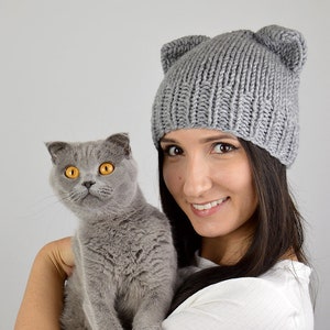 May include: A woman wearing a gray knitted beanie with cat ears holds a gray cat in her arms. The cat has bright yellow eyes.