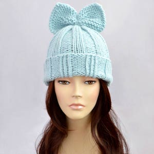 May include: A light blue knitted beanie with a large bow on top. The beanie is made of a soft, warm yarn and is perfect for keeping your head warm in the winter.