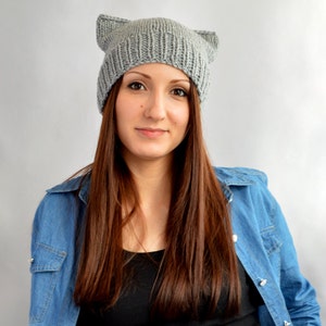 May include: A woman with long brown hair is wearing a gray knitted beanie with cat ears. She is also wearing a blue denim shirt.