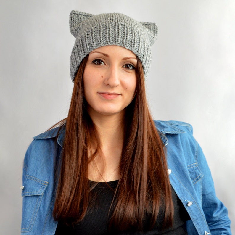 May include: A woman with long brown hair is wearing a gray knitted beanie with cat ears. She is also wearing a blue denim shirt.