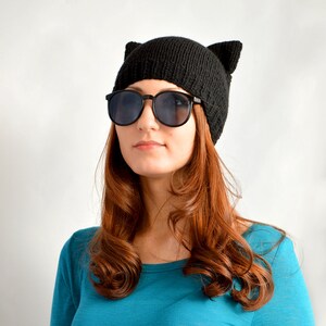 May include: A black knit cat ear beanie paired with black sunglasses. The beanie features two upright ears. The person is wearing a teal long-sleeve shirt. The background is plain white.