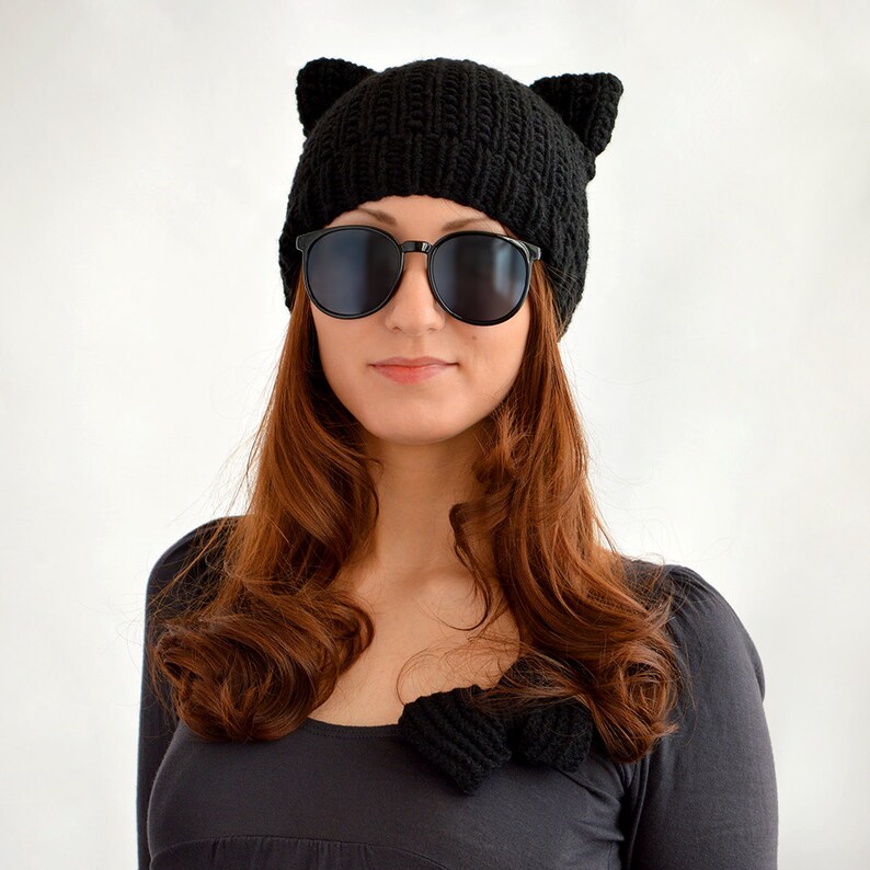 May include: A woman wearing a black knit beanie with cat ears and black sunglasses. The beanie is knitted with a textured pattern. The woman has long brown hair.