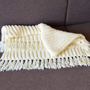 May include: A white crocheted blanket with fringe on the edges. The blanket is folded in half and has a textured pattern.