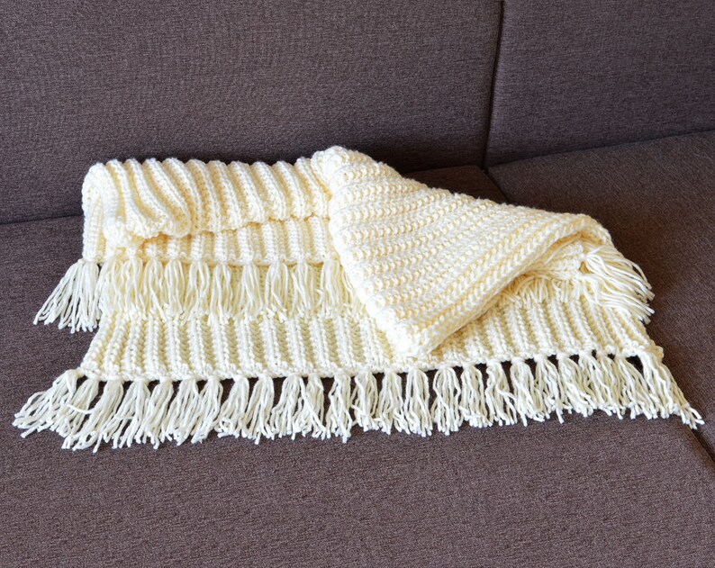 May include: A white crocheted blanket with fringe on the edges. The blanket is folded in half and has a textured pattern.