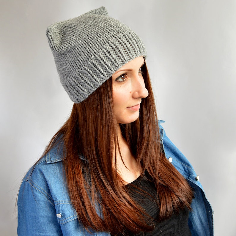 May include: A gray knitted beanie with cat ears. The beanie is worn by a person with long brown hair and a denim jacket.