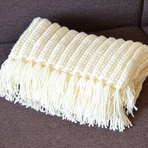 May include: A white crocheted throw blanket with fringe on the edges. The blanket is folded and lying on a brown couch.