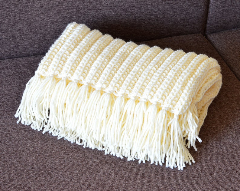 May include: A white crocheted throw blanket with fringe on the edges. The blanket is folded and lying on a brown couch.