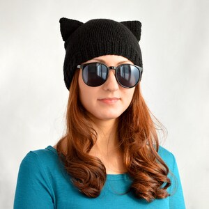 May include: A black knit cat ear beanie with a ribbed texture. The beanie features two upright ears at the top. The person is wearing black sunglasses and a teal long-sleeve shirt. The background is plain white.