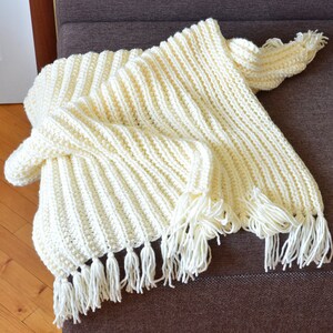May include: A cream-colored crocheted throw blanket with fringe on the edges. The blanket is folded and draped over a brown couch.