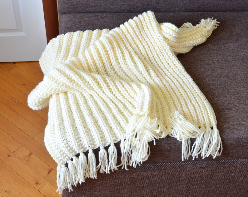 May include: A cream-colored crocheted throw blanket with fringe on the edges. The blanket is folded and draped over a brown couch.