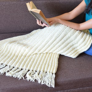 May include: A white crocheted throw blanket with fringe on a brown couch. The blanket is draped over the couch and a person is sitting on the couch reading a book.