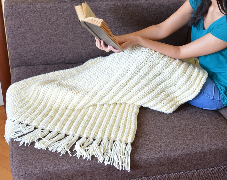 May include: A white crocheted throw blanket with fringe on a brown couch. The blanket is draped over the couch and a person is sitting on the couch reading a book.