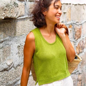 May include: A sleeveless, lime green knit top with a scoop neckline. The top is paired with white shorts. A straw basket is carried over the shoulder. The background is a stone wall.