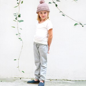 May include: A young person wearing a light pink beanie with a pom pom on top, a white t-shirt, gray sweatpants, and dark blue slip-on shoes.