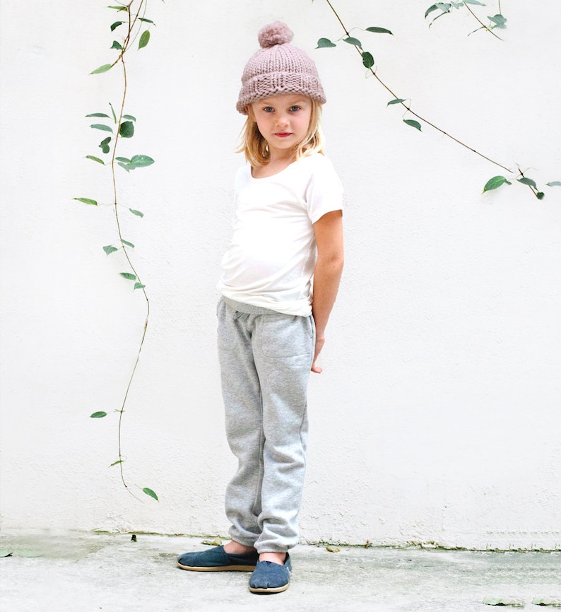 May include: A young person wearing a light pink beanie with a pom pom on top, a white t-shirt, gray sweatpants, and dark blue slip-on shoes.