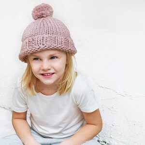 May include: A young person wearing a pink knitted beanie with a fluffy pom pom on top. The beanie is pulled down over their forehead and they are looking to the side with a smile.