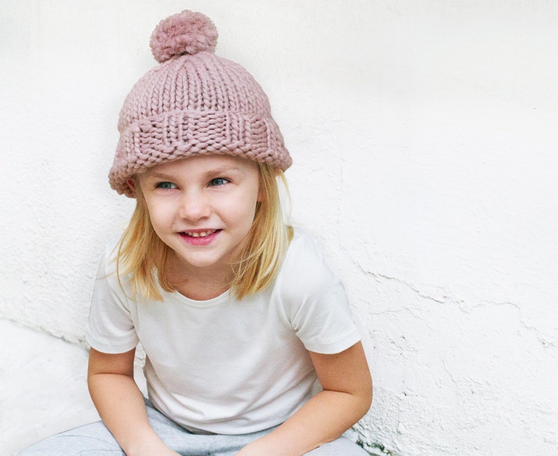 May include: A young person wearing a pink knitted beanie with a fluffy pom pom on top. The beanie is pulled down over their forehead and they are looking to the side with a smile.