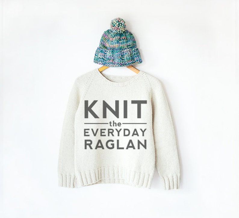 Op de afbeelding: Een witte gebreide trui met de tekst "KNIT the EVERYDAY RAGLAN" op de voorkant. De trui hangt aan een hanger en een kleurrijke gebreide muts ligt bovenop de hanger.