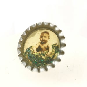 May include: A silver bottle cap with a picture of a baby inside. The picture is surrounded by green glitter.