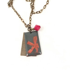 May include: A necklace with a dark gray rectangular pendant featuring a red abstract design. The pendant is attached to a brass chain with a small red crystal charm.
