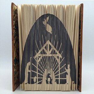 May include: A book art sculpture depicting the nativity scene. The book is folded into a three-dimensional shape, creating a silhouette of a stable with a star above it. The figures of Mary, Joseph, and the baby Jesus are visible inside the stable.