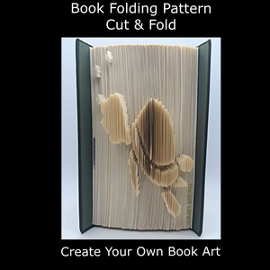 May include: An open book with pages folded to create a detailed bee design. The book has a dark green cover and the text "Book Folding Pattern Cut & Fold" and "Create Your Own Book Art" is displayed.
