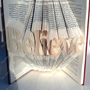 May include: A book sculpture with the word "BELIEVE" carved into the pages. The pages are fanned out to create a unique, artistic display. The book has a red cover and the text is in a serif font.