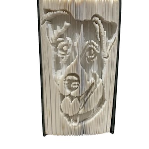 May include: A handcrafted book sculpture with a dog's face design. The pages are intricately folded and cut to reveal the dog's features. The book has a dark green spine, and the paper is a light cream colour, creating a striking contrast.