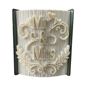Peut inclure: Livre décoratif avec les mots « Mr & Mrs » découpés dans les pages. Les lettres et les éléments décoratifs sont de couleur crème, sur fond de pages blanches. La tranche du livre est vert foncé.
