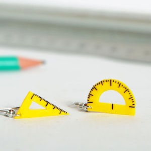 Peut inclure: Une paire de boucles d'oreilles en plastique jaune en forme de rapporteur et de triangle. Les boucles d'oreilles ont des crochets argentés.