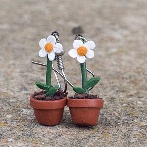 Puede incluir: Pendientes de margaritas hechos a mano. Cada pendiente presenta una margarita blanca con centro naranja, tallo y hojas verdes, y una pequeña maceta marrón. Los pendientes tienen ganchos de metal plateado.