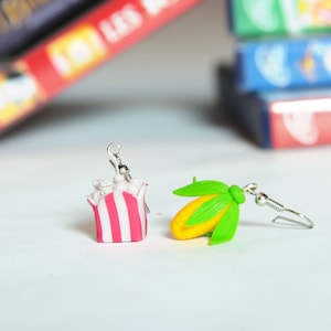 Peut inclure: Une paire de boucles d'oreilles faites à la main représentant un seau de pop-corn et un épi de maïs. Le seau de pop-corn est rayé de rose et de blanc avec du pop-corn blanc à l'intérieur. L'épi de maïs est jaune avec des feuilles vertes. Les boucles d'oreilles sont en pâte polymère et en fils d'oreille argentés.