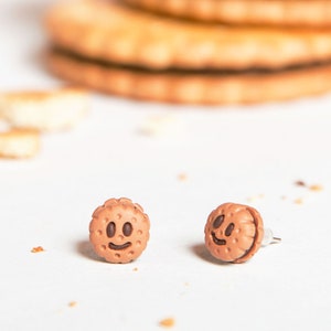 Puede incluir: Un par de pendientes de galleta marrón y blanco con un diseño de cara sonriente. Los pendientes están hechos de arcilla polimérica y tienen un cierre de poste.