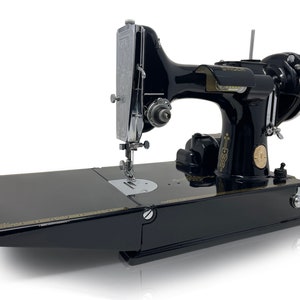 May include: A vintage black Singer sewing machine with chrome detailing. The machine has a flatbed design and gold trim. The Singer logo is visible. The machine is set against a white background. A classic sewing machine.