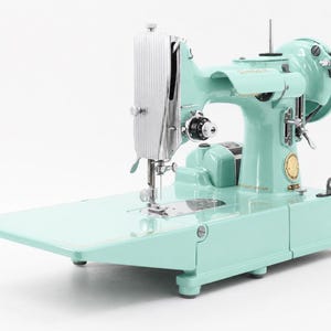 以下が含まれることがあります： ミントグリーンのSinger製ミシン。リブ付きのシルバーのフェイスプレート、側面の円形ゴールドエンブレム、フラットなベッドエクステンションが特徴です。ハンドホイール、針アセンブリ、およびさまざまな金属製の調整ノブとレバーを備えています。