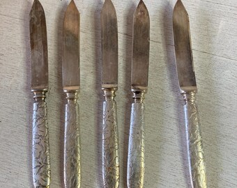May include: Set of five silver butter knives with ornate handles. The knives have a pointed tip and a smooth blade.